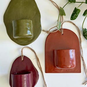 May include: Three wall-mounted ceramic planters with glossy finishes in olive green, terracotta orange, and deep burgundy. Each planter features a flat backplate with a hole for hanging and a cylindrical pocket, suspended by natural jute twine.