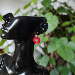 Può includere: Orecchini rossi circolari con cerchi dorati. Sono esposti su una testa di manichino nera, con uno sfondo di fogliame verde. Gli orecchini hanno una finitura lucida e un design minimalista.