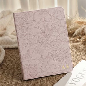 Puede incluir: Una funda para tableta de color rosa claro con un diseño floral en relieve. La funda presenta un patrón detallado de flores y hojas. Las iniciales doradas "A.E" son visibles en la esquina inferior derecha. La funda está sobre una superficie texturizada junto a una revista.