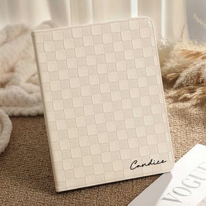 Puede incluir: Funda de tableta de color crema con un patrón de cuadros. La funda tiene el nombre "Candice" en cursiva. La funda está sobre una superficie marrón junto a una revista.