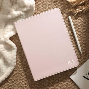May include: A light pink tablet case with the initials "B.A" in white. A white stylus and a small white and black book are next to the case. The case is on a brown surface with a white fluffy blanket and dried plants.