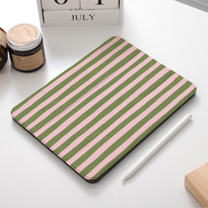 Pode incluir: Uma capa para tablet com um padrão de listras verdes e rosa. Uma caneta stylus branca repousa ao lado da capa. Dois frascos de vidro castanho com rótulos e um calendário de blocos de madeira mostrando julho também são visíveis.