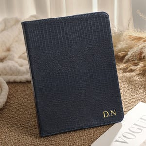 Puede incluir: Un cuaderno azul oscuro con un patrón de textura de piel de cocodrilo en la cubierta. En la esquina inferior derecha presenta las iniciales D.N. en color dorado. El cuaderno es rectangular con bordes cosidos y un aspecto limpio.