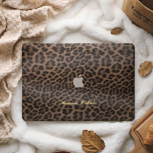 May include: A rectangular laptop cover featuring a brown and black leopard print pattern with a central silver Apple logo cutout and the gold text Monroe Wakutz printed in a cursive font on the lower portion of the surface.