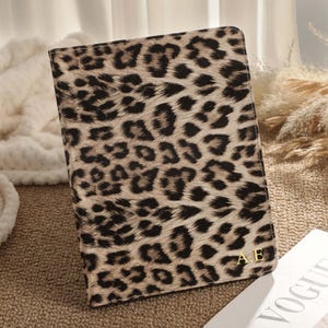 Puede incluir: Un portapasaporte rectangular con estampado de leopardo. La cubierta presenta un fondo beige con manchas negras y marrones. Las iniciales doradas "AE" son visibles en la esquina inferior derecha. También se ve una revista con la palabra "VOGUE".