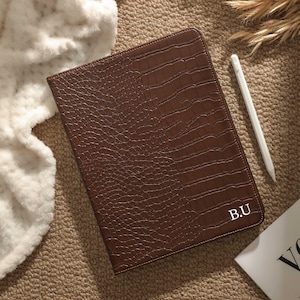 May include: A brown, faux-leather tablet case with a crocodile skin texture. The case has the initials "B.U." in white on the lower right corner. A white stylus and a book are next to the case. The case is on a brown surface.