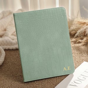 May include: A light green tablet case with a crocodile skin texture. The case has the gold initials "A.E" in the bottom right corner. A copy of Vogue magazine is visible in the background.