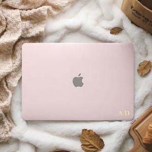 May include: A light pink laptop cover featuring a centered silver Apple logo and the gold-colored initials A.D. printed in the bottom right corner. The cover has a smooth, matte texture and is displayed from a top-down perspective.