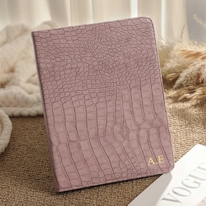 May include: A mauve-colored, faux-leather tablet case with a crocodile skin texture. The case has gold-colored initials "A.E" in the lower right corner. The case is resting on a textured surface with a magazine in the background.