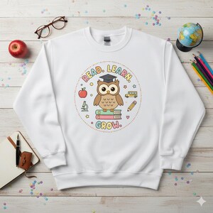 May include: White sweatshirt with a colorful graphic of a cartoon owl wearing a graduation cap, sitting on books, and surrounded by school-themed illustrations. The text "READ. LEARN. GROW." is also included in the design. Perfect for students and teachers.