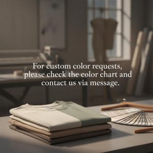May include: A stack of folded t-shirts in neutral colors, including beige, tan, and olive green, are displayed on a table. The text "For custom color requests, please check the color chart and contact us via message" is visible. A color chart and hangers are also present.