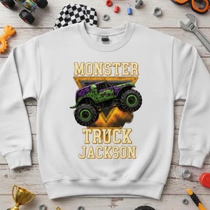 May include: White sweatshirt featuring a monster truck graphic with the text "MONSTER TRUCK JACKSON". The monster truck is purple and green with large black tires. The design is set against a yellow triangle.