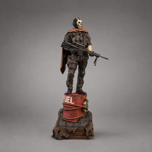 May include: A detailed figurine of a soldier wearing a skull mask, tactical gear, and a brown cape, holding a rifle. The figure stands atop a weathered red fuel barrel marked with the word FUEL, wrapped in rope, resting on a dark textured base.