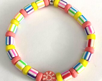 May include: A colorful bracelet featuring a mix of cylindrical beads in yellow, pink, and white with striped patterns in various colors. The bracelet includes a round charm with a sun design and the text "BJFDESIGNS".