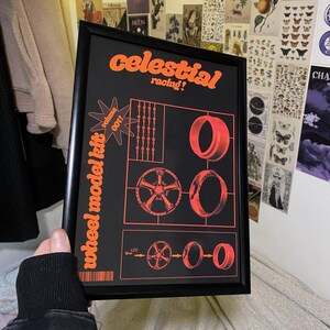Puede incluir: Póster enmarcado en negro con texto y gráficos en naranja. El póster dice "celestial racing!" y "wheel model kit volume 001!". Presenta ilustraciones de ruedas y neumáticos de coche. El póster lo sostiene una persona.