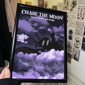 Pode incluir: Uma impressão artística emoldurada com o texto "CHASE THE MOON" e "RACE AMONG THE STARS". A obra apresenta uma paisagem roxa e preta com nuvens e uma lua cheia. A moldura é preta.