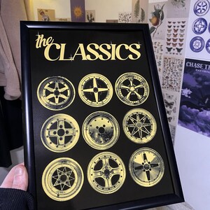 Puede incluir: Impresión enmarcada en negro con el texto "the CLASSICS" en dorado sobre nueve diseños de llantas de coche doradas. La impresión está sujeta por una mano.