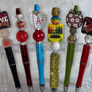 May include: A collection of decorative pens with various toppers, including an apple, a heart, and a crayon design. Some pens have the text "TEACH LOVE INSPIRE". The pens have black, red, blue, and green barrels, and are displayed in a clear holder.
