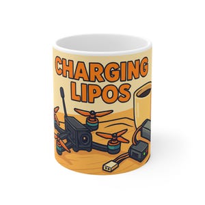 以下が含まれることがあります： オレンジ色のブロック文字で「CHARGING LIPOS」と書かれた白いセラミックマグ。マグカップには、ドローン、コーヒーカップ、充電機器の漫画イラストがベージュの背景に描かれています。