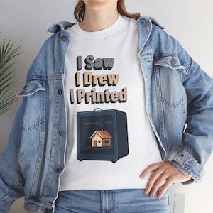 Op de afbeelding: Wit T-shirt met de tekst "I Saw I Drew I Printed" in een gelaagde lettertype. Onder de tekst staat een afbeelding van een 3D-printer met een klein huisje. Het shirt wordt gedragen met een blauw spijkerjasje en jeans.