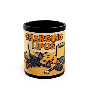 Peut inclure: Mug en céramique noire et beige avec l'inscription orange "CHARGING LIPOS". L'illustration représente un drone, une tasse de café et du matériel de chargement. L'intérieur et le bord de la tasse sont noirs.