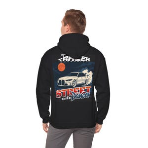 Könnte beinhalten: Schwarzer Kapuzenpullover mit einer Grafik eines weißen Sportwagens, der driftet, mit dem Text "STREET DANCER" und japanischen Schriftzeichen. Das Design beinhaltet einen roten Mond und die Worte "RACER COMMUNITY".