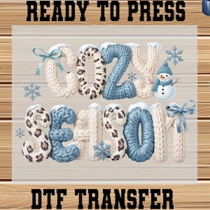 May include: A DTF transfer design with the words "COZY SEASON" in a knitted font, with blue and cream colors, snowflakes, and a snowman. The text "READY TO PRESS" and "DTF TRANSFER" are at the top and bottom.