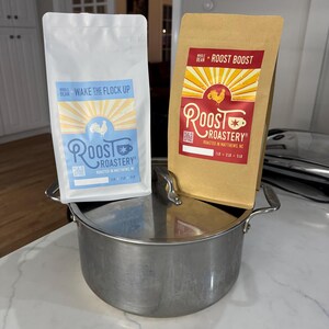 May include: Two bags of whole bean coffee from Roost Roastery. One bag is white with the text "Wake the Flock Up" and the other is brown with the text "Roost Boost". Both bags have a rooster logo and are sitting on a stainless steel pot.