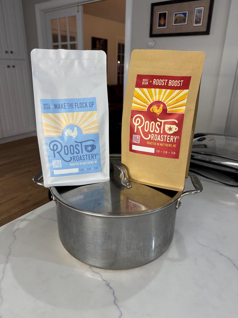 May include: Two bags of whole bean coffee from Roost Roastery. One bag is white with the text "Wake the Flock Up" and the other is brown with the text "Roost Boost". Both bags have a rooster logo and are sitting on a stainless steel pot.