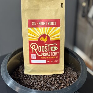 May include: A tan coffee bag with the text "Roost Boost" and a red and yellow rooster logo. The bag is in a black container filled with coffee beans. The bag states "Roasted in Matthews, NC".