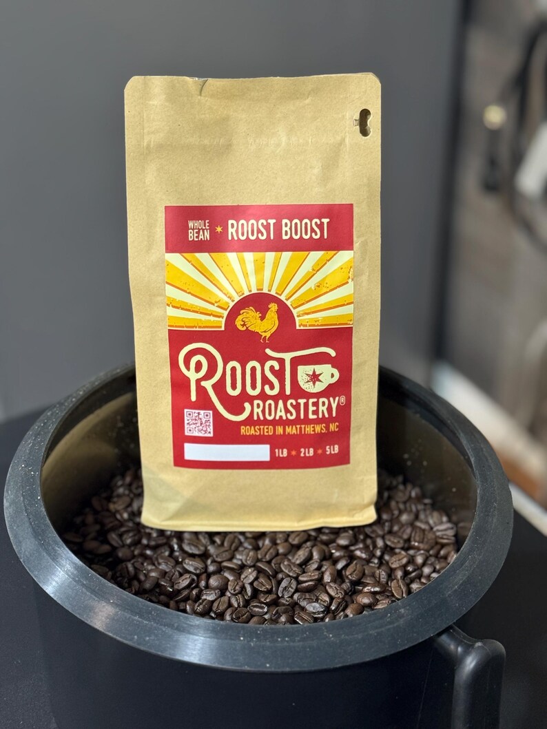 May include: A tan coffee bag with the text "Roost Boost" and a red and yellow rooster logo. The bag is in a black container filled with coffee beans. The bag states "Roasted in Matthews, NC".