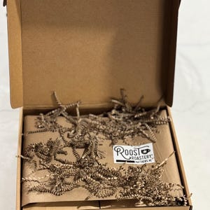May include: A brown cardboard box, open to reveal a package wrapped in brown paper and cushioned with shredded brown paper. A white label with the text "Roost Roastery" is visible on the package.
