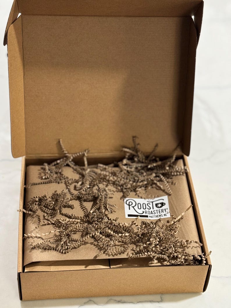 May include: A brown cardboard box, open to reveal a package wrapped in brown paper and cushioned with shredded brown paper. A white label with the text "Roost Roastery" is visible on the package.