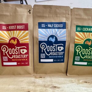 May include: Five brown paper coffee bags from Roost Roastery. Each bag has a different label design with the Roost Roastery logo, a rooster graphic, and text such as "Roost Boost" and "Wake the Flock Up". The bags contain whole bean coffee.