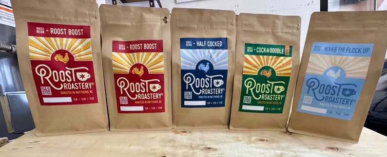 May include: Five brown paper coffee bags from Roost Roastery. Each bag has a different label design with the Roost Roastery logo, a rooster graphic, and text such as "Roost Boost" and "Wake the Flock Up". The bags contain whole bean coffee.