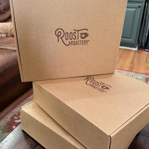 May include: Three stacked, unopened cardboard boxes. The boxes are a light brown colour and have the "Roost Roastery" logo printed in brown on the top of each box. The boxes are on a dark brown wooden table.