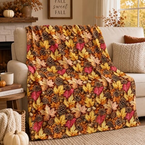 May include: A soft throw blanket featuring a repeating autumn pattern of yellow, orange, and pink maple leaves interspersed with brown pinecones and acorns. The fabric displays a dense, colorful botanical print covering the entire surface.