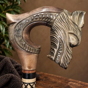 May include: A wooden walking stick handle carved into the shape of a wolf head with detailed fur texture. The handle features a smooth, light brown finish, while the shaft below includes a black and light wood section with a cross-hatch pattern.