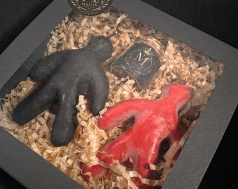 May include: A black gift box with a clear window containing a black figure, a red figure, and a small glass jar with a black label featuring a gold crown and the letter M, all nestled on a bed of light brown shredded paper filler material.