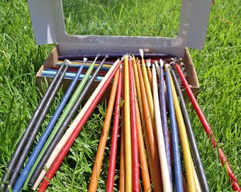 May include: A collection of thin, multi-colored taper candles in shades of red, orange, yellow, blue, green, purple, and white, arranged in a fan pattern extending from an open cardboard box with a clear plastic window on the lid.