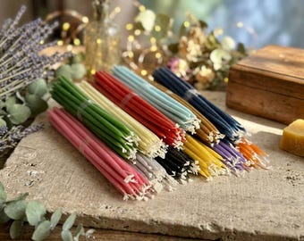 May include: A collection of thin, tapered candles bundled together in various colors including pink, green, cream, red, light blue, dark blue, black, yellow, purple, and orange, each featuring a visible white cotton wick at the top.