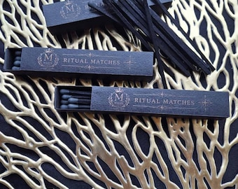 May include: Three black rectangular matchboxes labeled RITUAL MATCHES with a crown and M logo. Each box contains black matches with dark gray tips. A cluster of loose black matches with dark gray tips is fanned out across the top of the boxes.