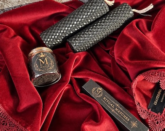 May include: Two black honeycomb-textured beeswax candles, a small glass jar with a black label reading Magik, and a black rectangular box labeled Ritual Matches are arranged on a deep red velvet fabric with a decorative lace border.
