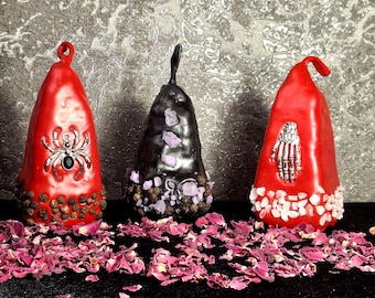 May include: Three cone-shaped ornaments with loops at the top. The left is red with a silver spider and dark peppercorns. The center is black with purple crystals and peppercorns. The right is red with a silver skeleton hand and pink crystals.
