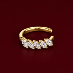 May include: A gold-toned ear cuff featuring five marquise-cut clear stones arranged in a row along the front curve. The metal band is smooth and polished, with an open design that allows for placement on the ear cartilage without a piercing.