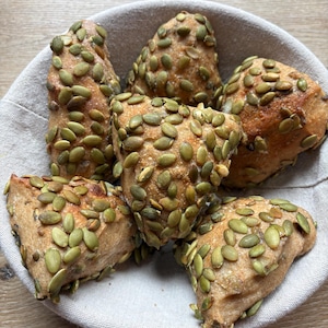 May include: A collection of five triangular-shaped bread rolls topped with green pumpkin seeds, arranged inside a round, light-colored fabric-lined basket. The rolls have a golden-brown crust and are densely covered in seeds.