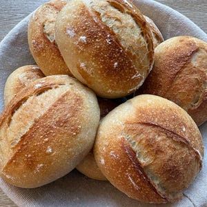 May include: A collection of round, golden-brown artisan bread rolls with a crisp, scored crust and a dusting of flour, arranged inside a round, light-colored fabric-lined basket. The rolls feature a distinct diagonal slash across the top.