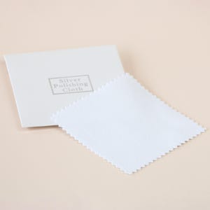 May include: A white silver polishing cloth with a zig-zag edge sits beside a white rectangular box. The box has the words "Silver Polishing Cloth" printed in grey.