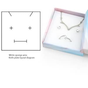Op de afbeelding: Een sieradenset in een decoratieve doos. De set bevat een zilveren ketting, oorbellen en een ring. De doos is wit met een pastelroze en blauw ontwerp. Links staat een diagram voor een mesplaatindeling.