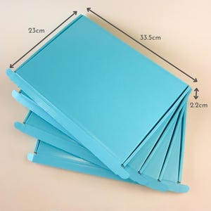 Peut inclure: Une pile de boîtes en carton bleu clair. Les boîtes sont rectangulaires, mesurant 33,5 cm de long, 23 cm de large et 2,2 cm de haut. Les boîtes sont soigneusement disposées, la boîte supérieure étant légèrement ouverte.
